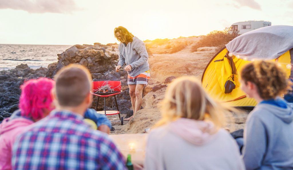group of friends camping on the beach, man preparing fire for barbecue